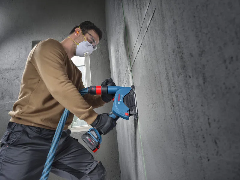 A person wearing safety equipment uses an angle grinder to cut a straight line in a concrete wall.