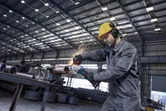 A person wearing safety equipment uses an angle grinder on a metal pipe, creating sparks.