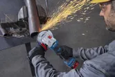 A person wearing safety equipment uses an angle grinder to cut a metal pipe.