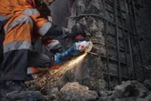 A person wearing safety equipment cuts concrete with an angle grinder, producing sparks.