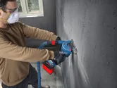 A person wearing safety equipment uses an angle grinder to cut a groove in a concrete wall.