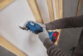 A person wearing safety equipment cuts a square opening in drywall using a multifunction tool.