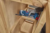 A person wearing safety equipment cuts a rectangular opening in a wooden cabinet using a multifunction tool.