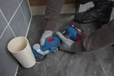A person wearing safety equipment uses a multifunction tool to cut floor tile next to a pipe.