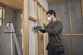 A person wearing safety equipment uses a multifunction tool to cut drywall.