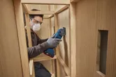 A person wearing safety equipment uses a multifunction tool to cut wood inside a cabinet.