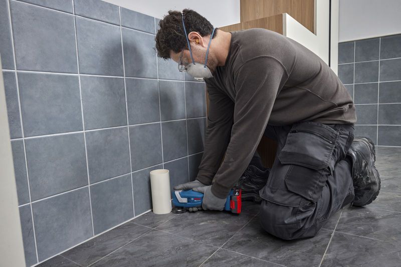 A person wearing safety equipment uses a multifunction tool to cut floor tiles.