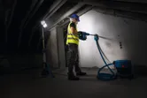 一名穿戴安全裝備的工人在無線工地照明燈下用電鑽鑽牆。
