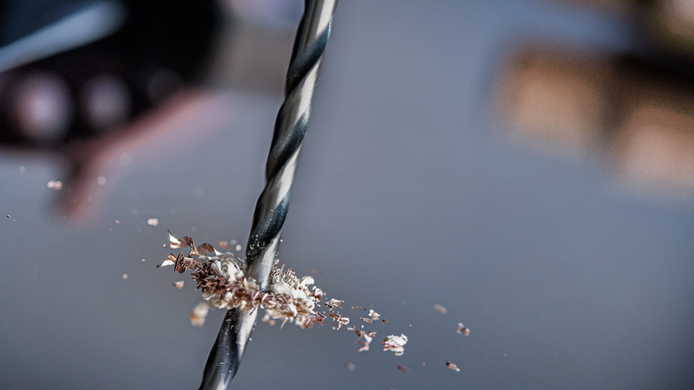鑽頭在金屬上鑽孔，金屬屑散落在周圍。