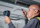 A person wearing safety equipment checks air duct temperature using a thermo detector.