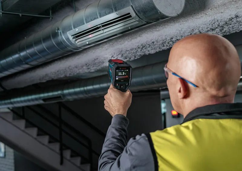 A person wearing safety equipment uses a thermo detector to check an air duct.