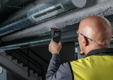 A person wearing safety equipment uses a thermo detector to check an air duct.