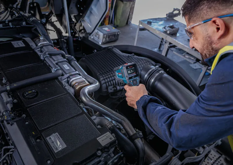 A person wearing safety equipment uses a thermo detector to check engine temperature.