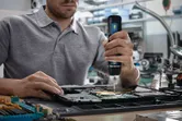 Person using a cordless screwdriver to assemble electronic components on a circuit board.