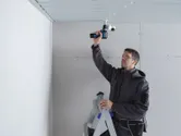 一名穿戴安全裝備的人員站在梯子上，使用無線電鑽在天花板上作業。