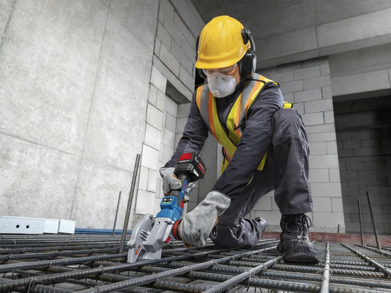 A worker wearing safety equipment uses a cordless rebar cutter on steel rods.