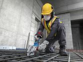 A worker wearing safety equipment uses a cordless rebar cutter on steel rods.