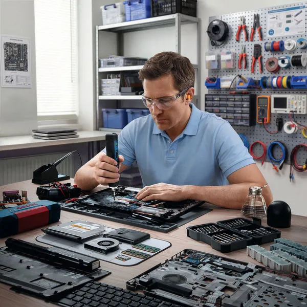 Person wearing safety equipment uses a cordless screwdriver to assemble a computer motherboard.