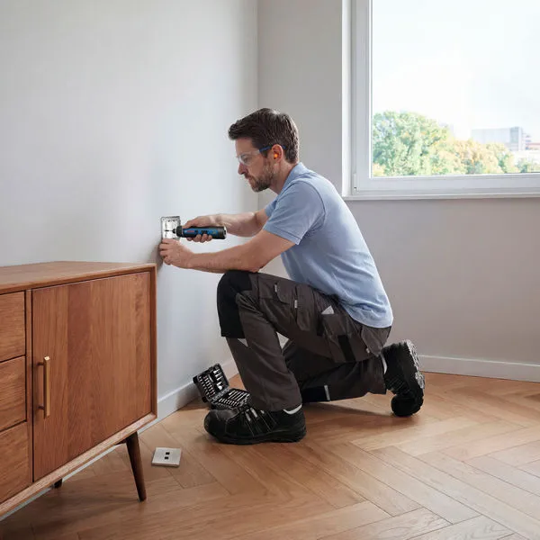 A person wearing safety equipment uses a cordless screwdriver on a wall outlet.