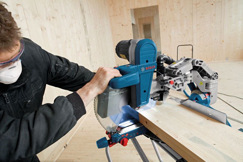A person wearing safety equipment cuts a wood plank with a sliding mitre saw.