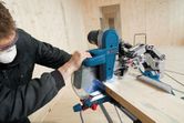 A person wearing safety equipment cuts a wood plank with a sliding mitre saw.