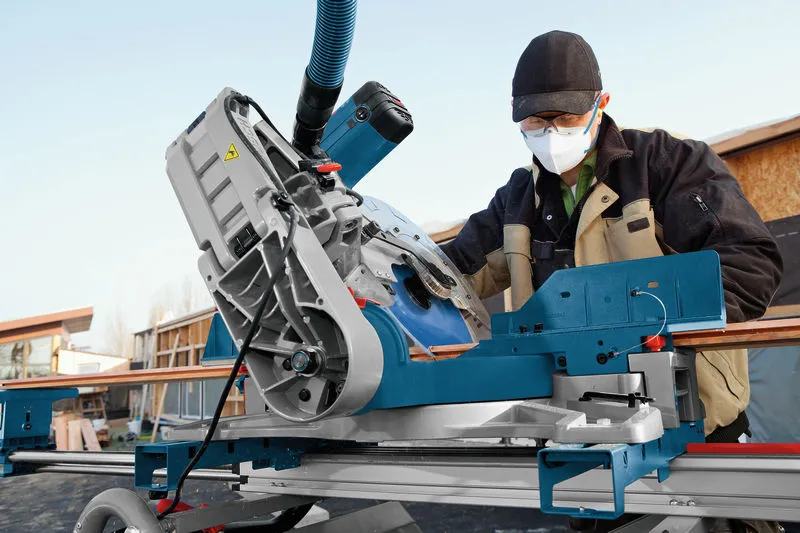 A person wearing safety equipment operates a mitre saw to cut wood on a jobsite.