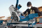 A person wearing safety equipment operates a mitre saw to cut wood on a jobsite.