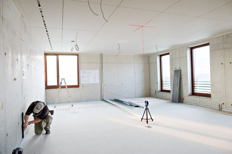 Person kneeling to measure a wall while laser leveling tool projects lines across a bright room.