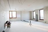 Person kneeling to measure a wall while laser leveling tool projects lines across a bright room.